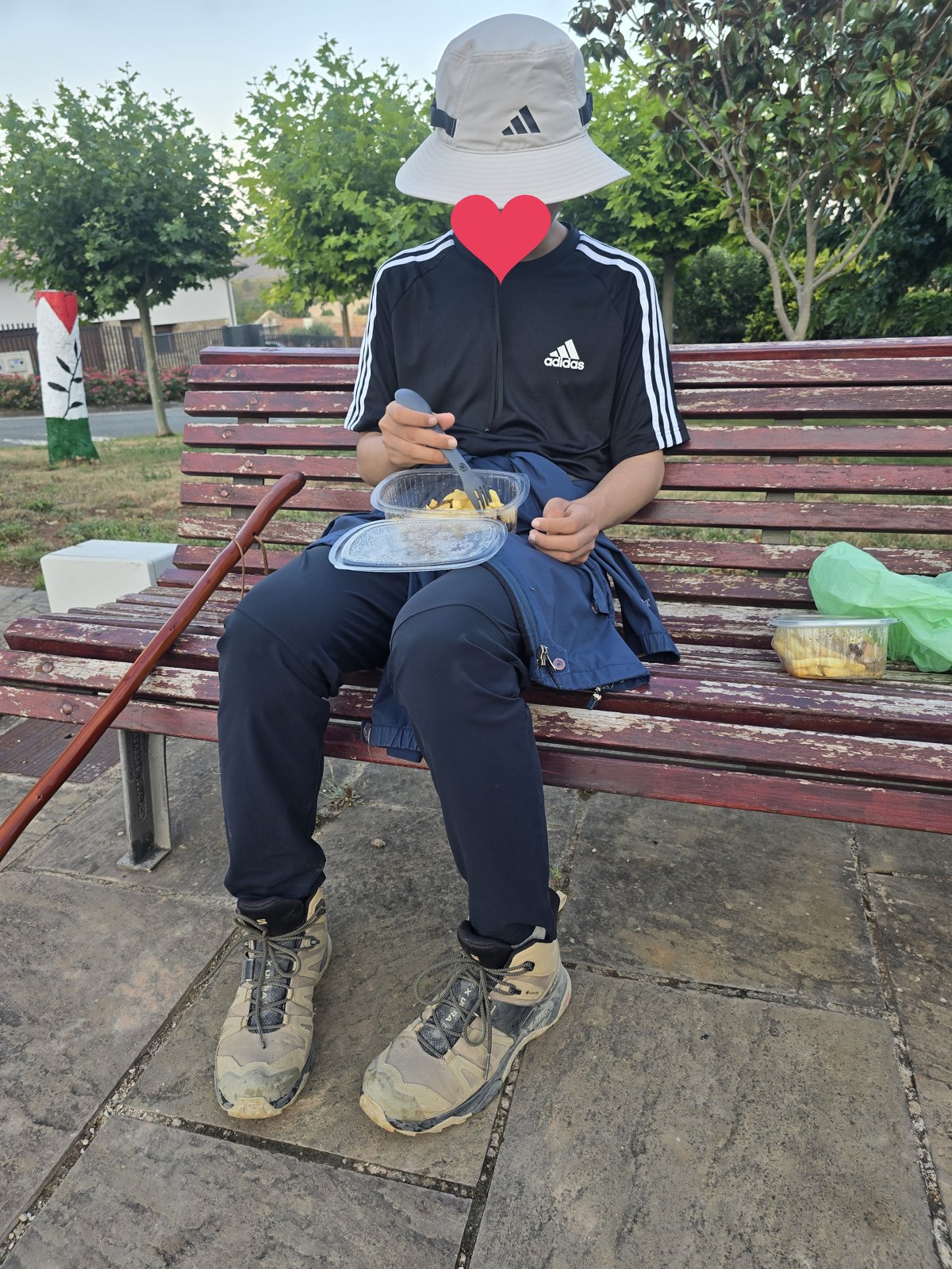 마을 벤치에 앉은 사람이 무릎에 플라스틱 도시락을 얹고 포크로 먹고 있다. 옆에 초록 비닐봉지와 반찬통이 놓여 있고 벤치 팔걸이에 붉은 나무 지팡이가 기대어 있다