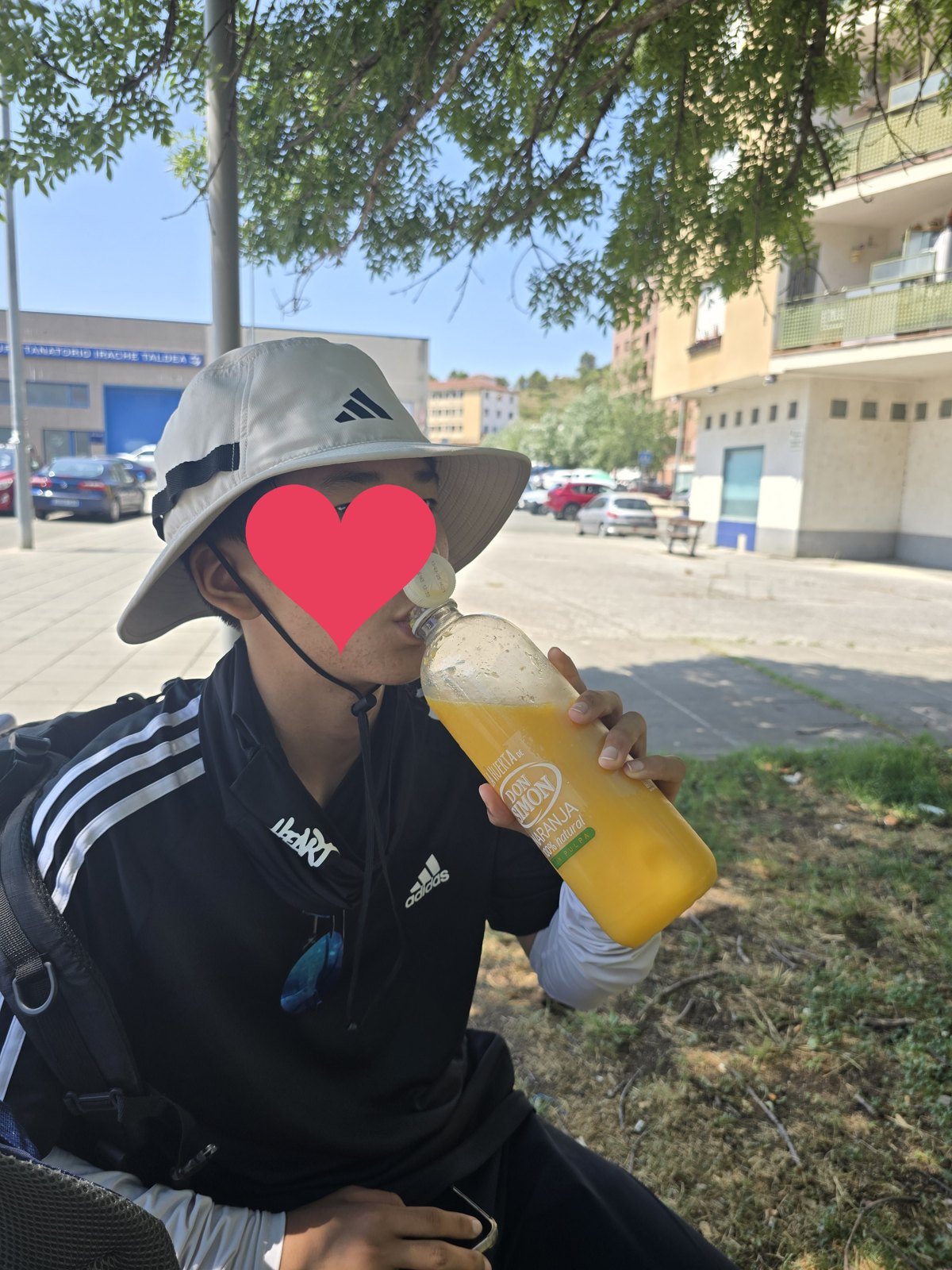 길가 나무 그늘에 앉은 사람이 오렌지주스 병을 들이켜고 있다. 뒤로 도로와 건물, 간판이 보인다