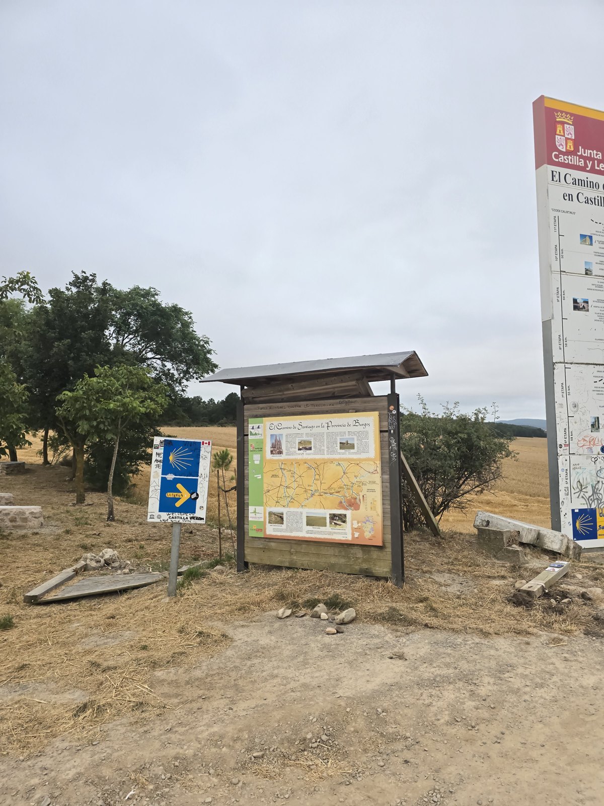 흙길 옆 풀밭에 나무 지붕을 얹은 안내판과 흰 세로 안내판이 나란히 서 있다. 뒤로 나무 몇 그루와 벤 밭이 보인다