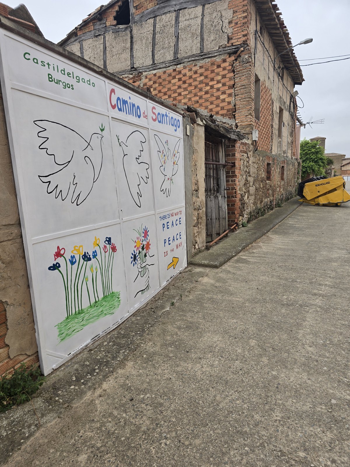 골목에 면한 흰 철문. 여섯 칸으로 나눠 비둘기 세 마리와 꽃, 꽃을 든 손을 그렸고 오른쪽 아래 칸에 파란 영어 글씨와 노란 화살표가 있다. 문 옆은 반목조 벽이 드러난 무너져 가는 집이다