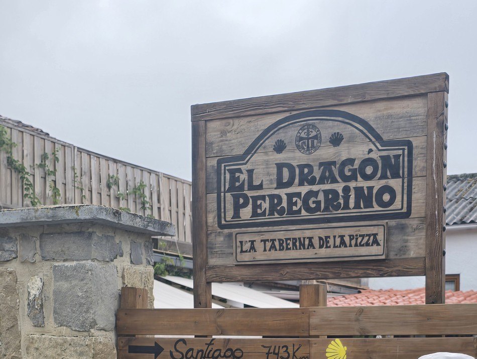 나무 간판에 EL DRAGON PEREGRINO, LA TABERNA DE LA PIZZA라고 새겨져 있고 그 아래 나무판에 산티아고 743km라고 손글씨로 적혀 있다