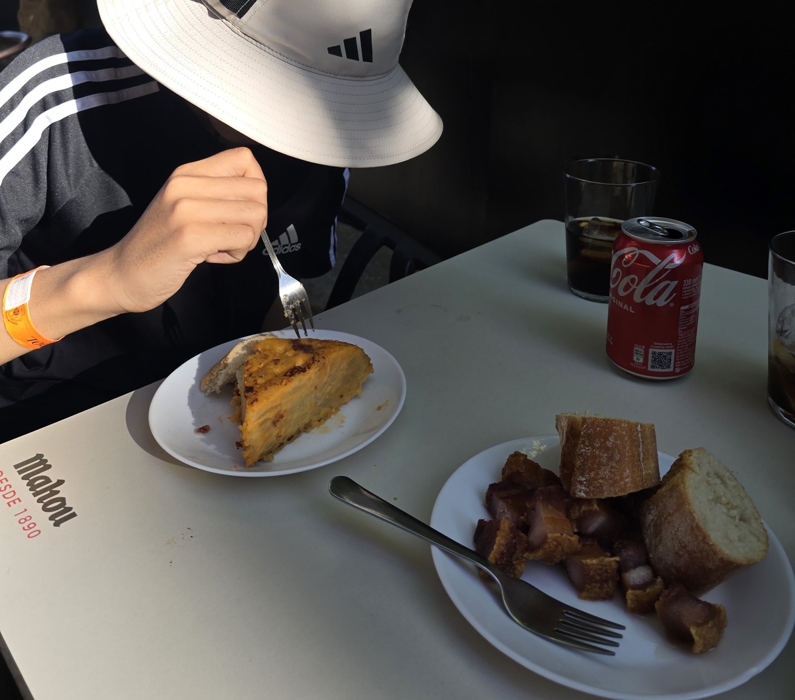 바 야외 탁자에 접시 두 개가 놓여 있다. 하나에는 두툼한 감자 오믈렛 한 조각과 빵이, 다른 하나에는 잘게 썬 고기와 바게트 조각이 담겨 있다. 옆에 콜라 캔과 잔이 있다