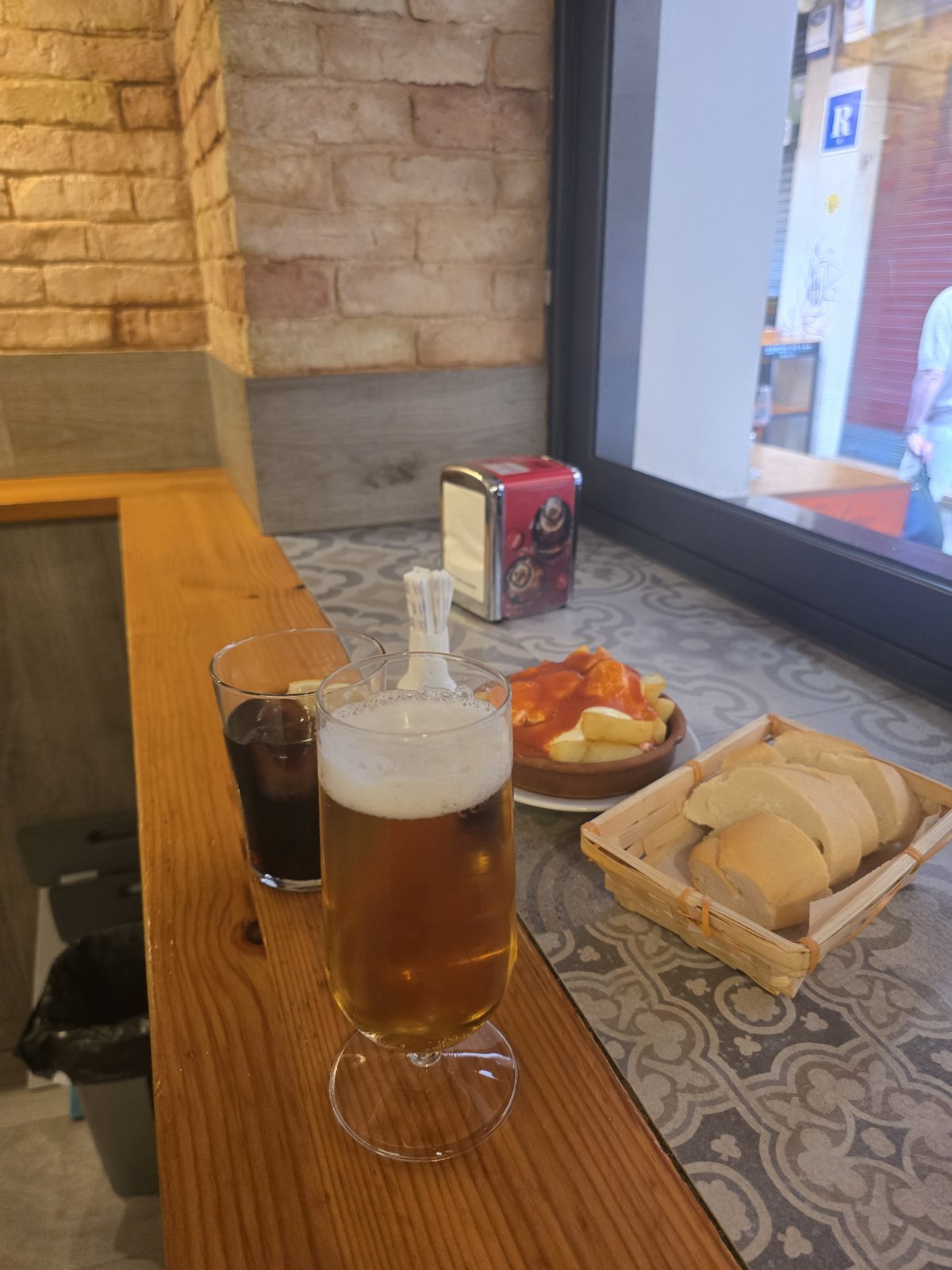 바 안쪽 나무 카운터에 맥주 한 잔과 콜라 잔이 놓여 있고, 붉은 소스를 끼얹은 감자 요리 접시와 빵을 담은 나무 바구니가 함께 있다