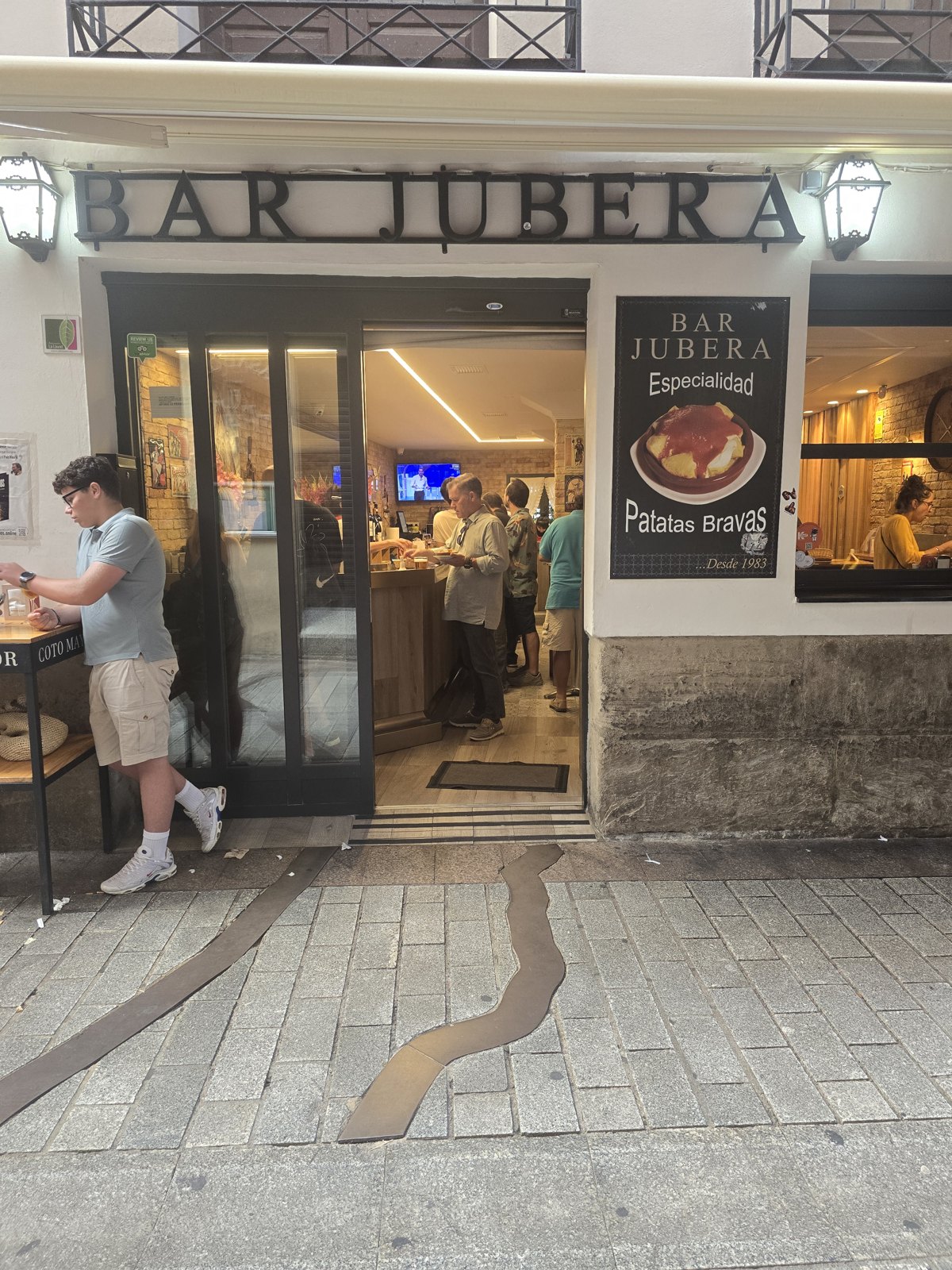 BAR JUBERA라고 쇠글자를 붙인 가게 앞. 오른쪽 벽에 감자 요리 사진과 Especialidad Patatas Bravas, Desde 1983이라고 적힌 검은 간판이 걸려 있다. 바닥 포석에 구불구불한 금속 띠가 박혀 있다