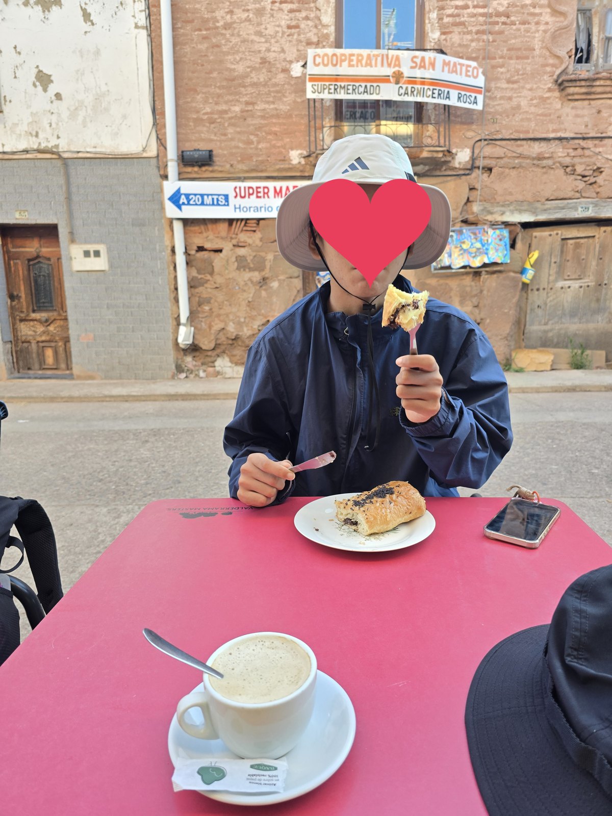 붉은 야외 탁자 위 흰 접시에 시금치를 넣어 구운 빵이 놓여 있고 검은깨가 뿌려져 있다. 앞쪽에 카페 콘 레체 한 잔이 있고 건너편 벽에 COOPERATIVA SAN MATEO라고 적힌 간판이 걸려 있다