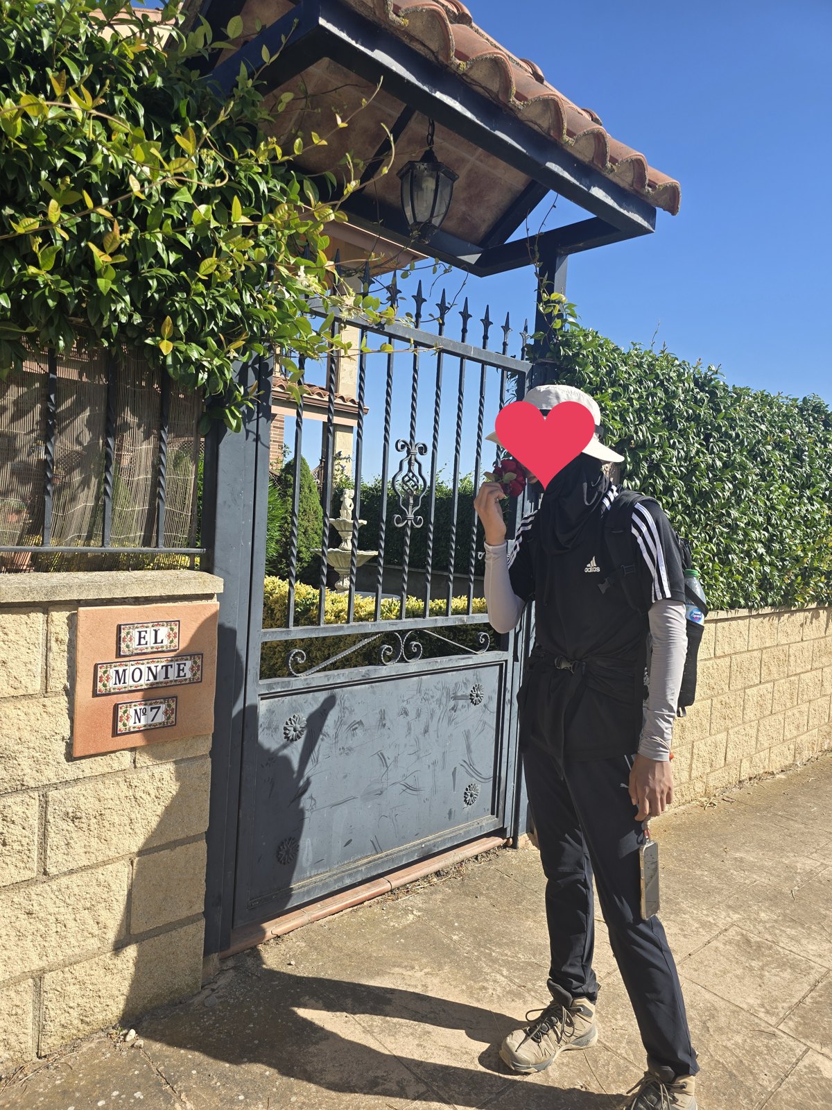 기와 지붕을 얹은 철제 대문. 문기둥에 EL MONTE Nº 7이라고 적은 타일 문패가 박혀 있고 철문 너머로 돌 분수와 손질한 정원이 보인다. 대문 옆에 배낭 멘 사람이 장미 한 송이를 들고 서 있다