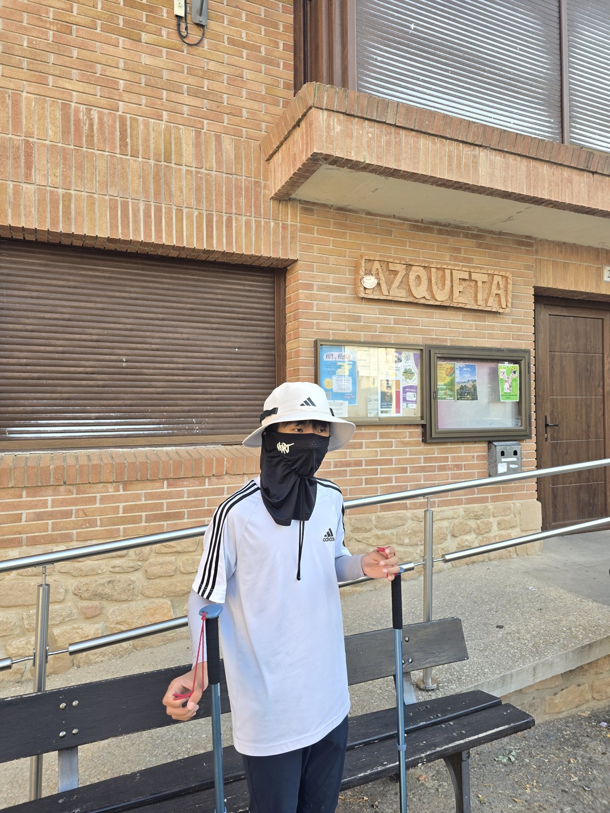 마을 건물 벽에 AZQUETA라고 적힌 간판과 유리 게시판이 붙어 있고 그 앞 벤치 옆에 넥게이터로 얼굴을 가린 사람이 스틱을 짚고 서 있다