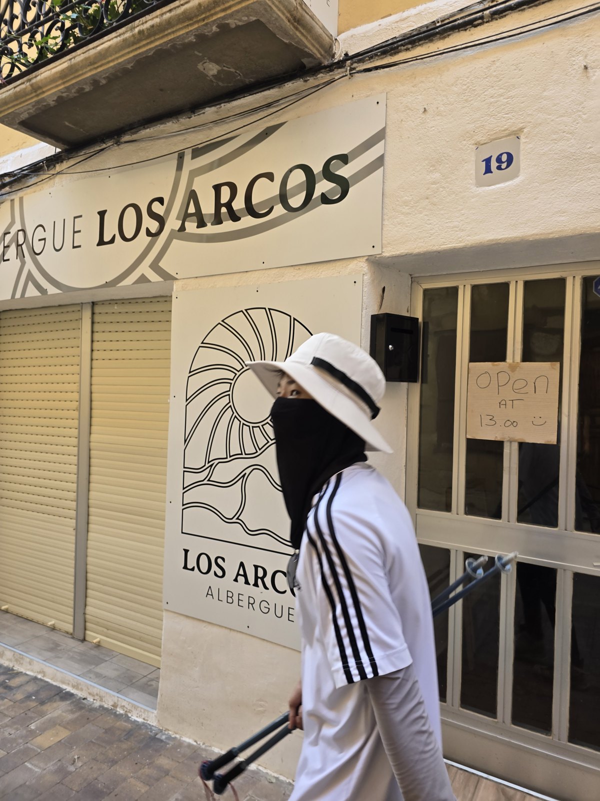흰 벽에 ALBERGUE LOS ARCOS라고 적힌 간판이 붙은 건물 앞을 넥게이터로 얼굴을 가린 사람이 지나간다. 문에 영업 시각을 적은 종이가 붙어 있다