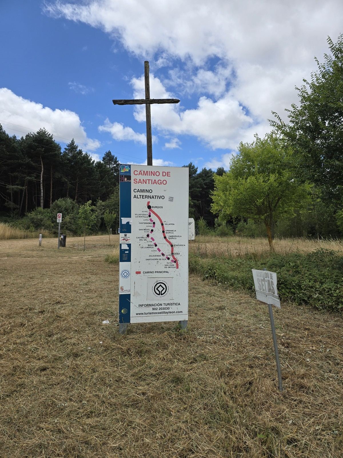 풀밭에 선 나무 십자가 옆에 CAMINO DE SANTIAGO 라고 적힌 파란 안내판이 있고 판에 붉은 선으로 그린 노선도가 인쇄돼 있다