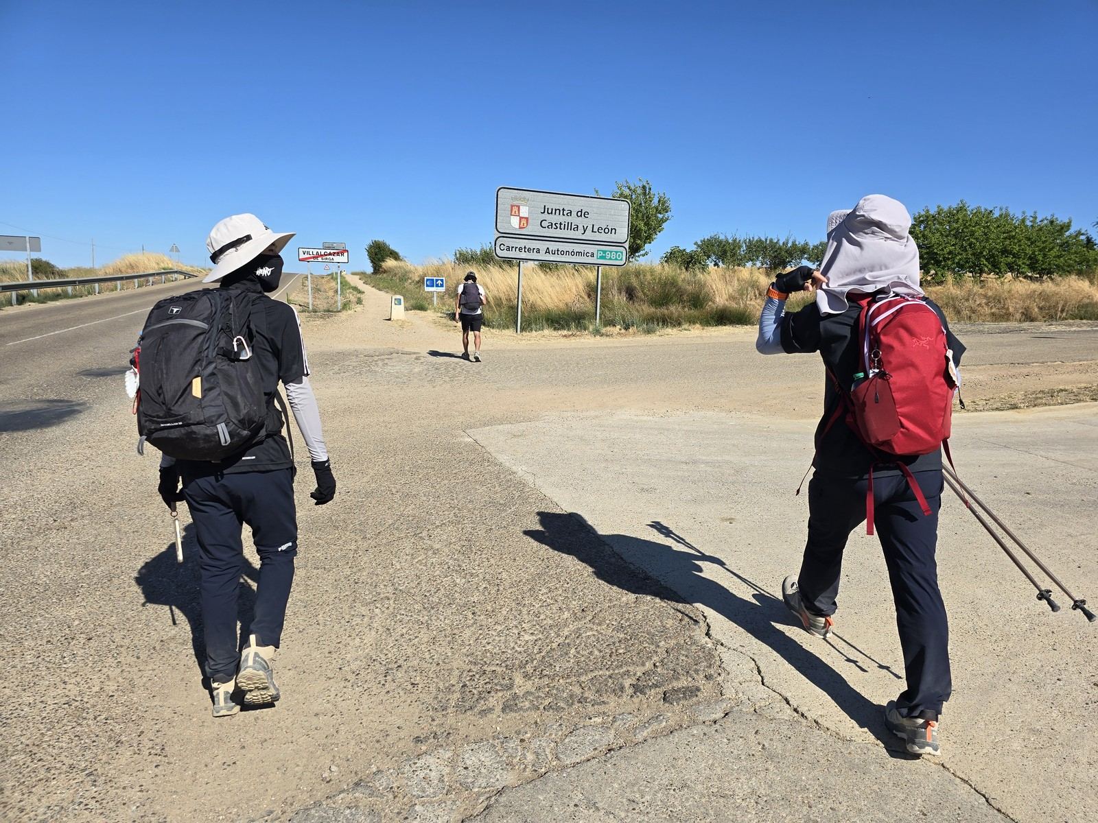 도로변 흰 표지판에 VILLALCÁZAR DE SIRGA 라고 적혀 있다. 뒤로 두 사람이 마을을 나서고 있다