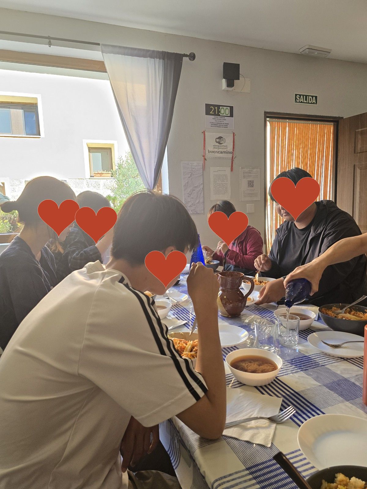 알베르게 식당의 긴 식탁에 순례자들이 마주 앉아 밥을 먹고 있다. 상에 국물 그릇과 면·밥 접시, 물병이 놓여 있고 벽에 21:00 식당 마감 안내가 붙어 있다. 앉은 사람들의 얼굴은 붉은 하트로 가려져 있다