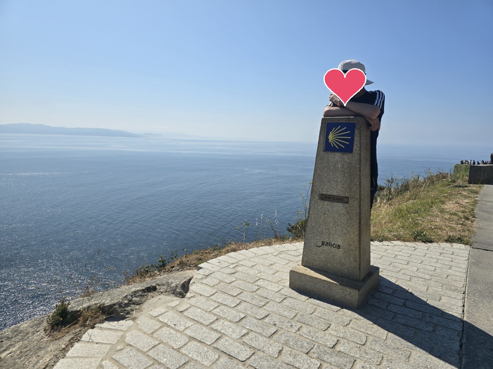 바다를 등지고 선 화강암 표석에 조가비 표식과 'Km 0,000'이 새겨져 있고 그 옆에 사람이 기대 서 있다