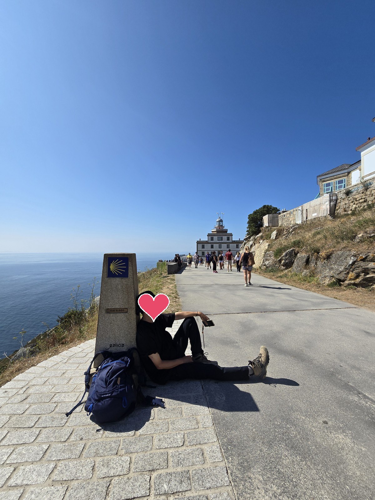 배낭을 내려놓고 Km 0.000 표석에 기대앉은 사람. 길 끝에 흰 등대 건물이 보인다