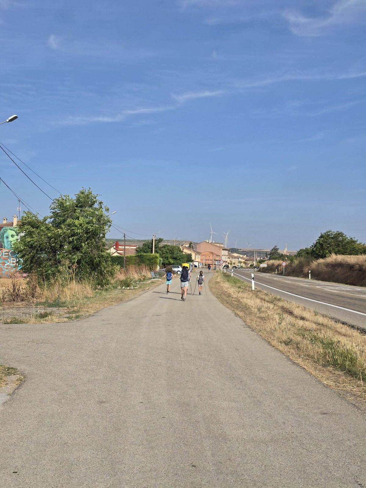 아침 햇살이 드는 아스팔트 도로. 양쪽에 마른 풀과 나무가 있고 길 끝에 마을 지붕이 보인다