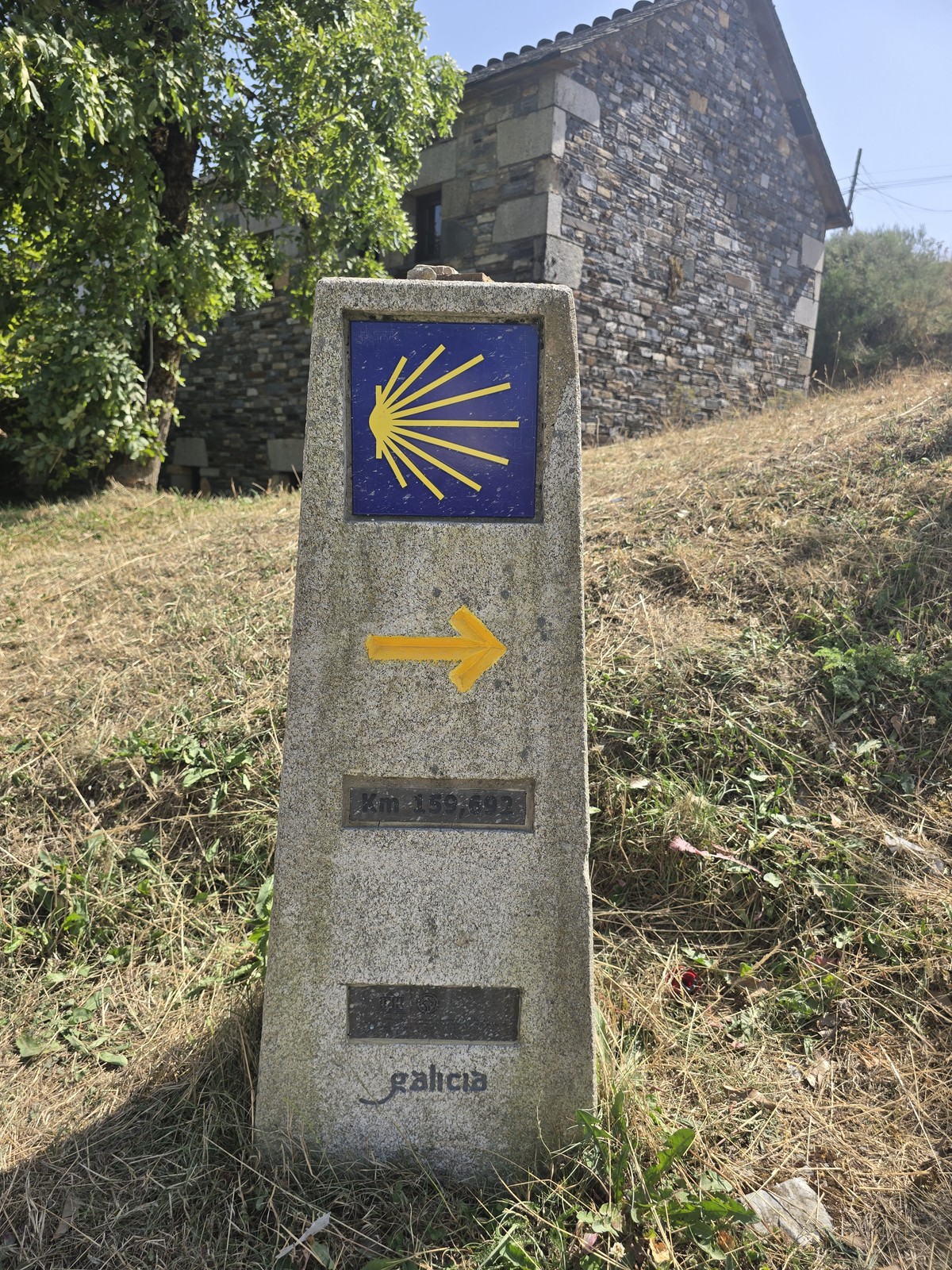 풀밭에 선 콘크리트 표석. 노란 조가비 표식과 화살표, 'Km 159,692', 'galicia' 글자가 새겨져 있다. 뒤로 돌집이 보인다