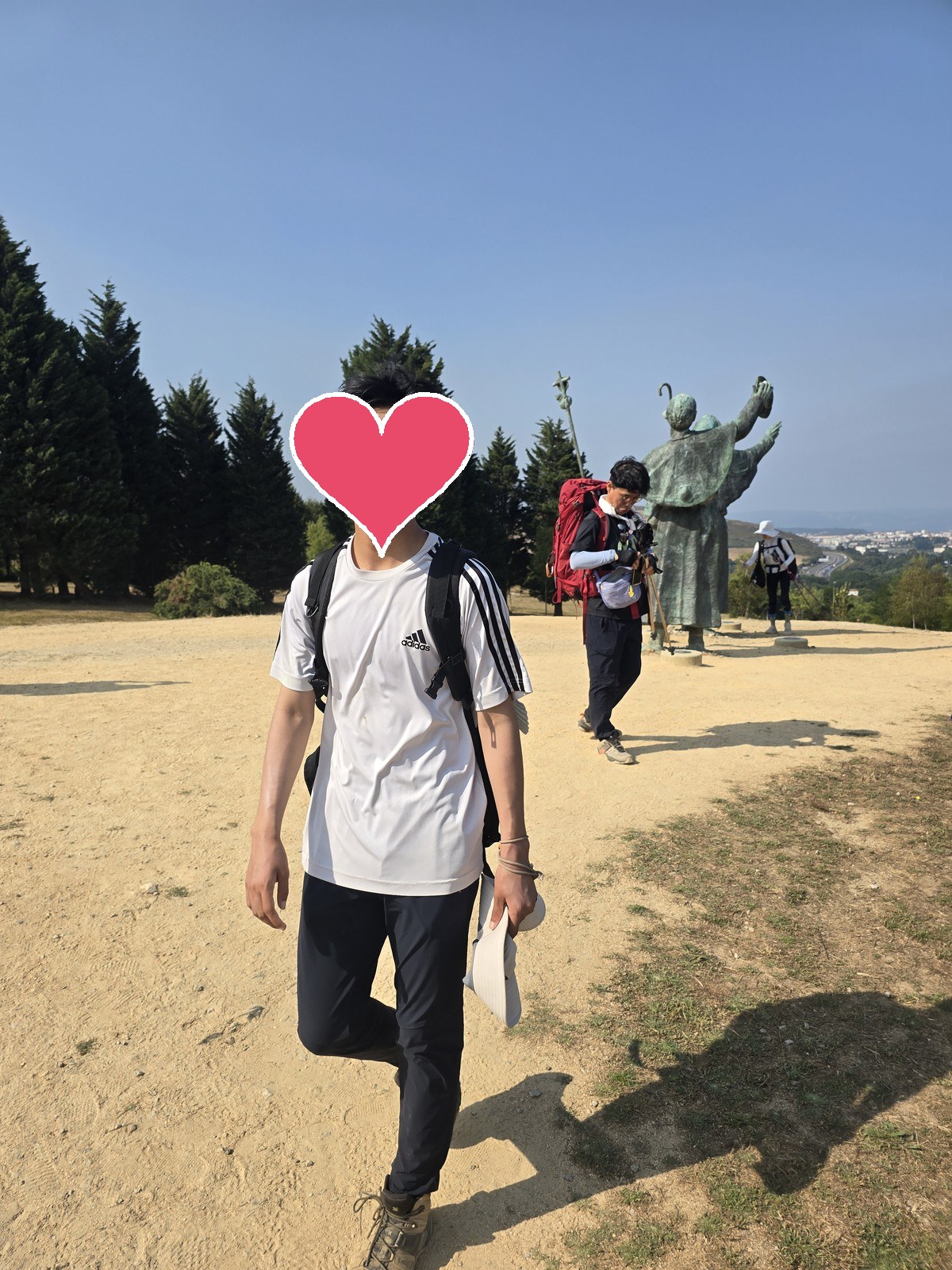 배낭 멘 순례자가 마른 흙길을 걸어오고 뒤로 청동 순례자상과 도시가 보인다